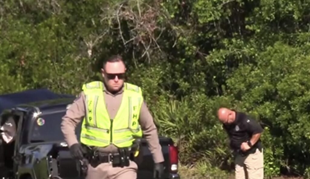 A police officer in a high-visibility vest stands near a vehicle at a crash scene on State Road 44 in DeLand, with another officer inspecting the ground amidst dense greenery.